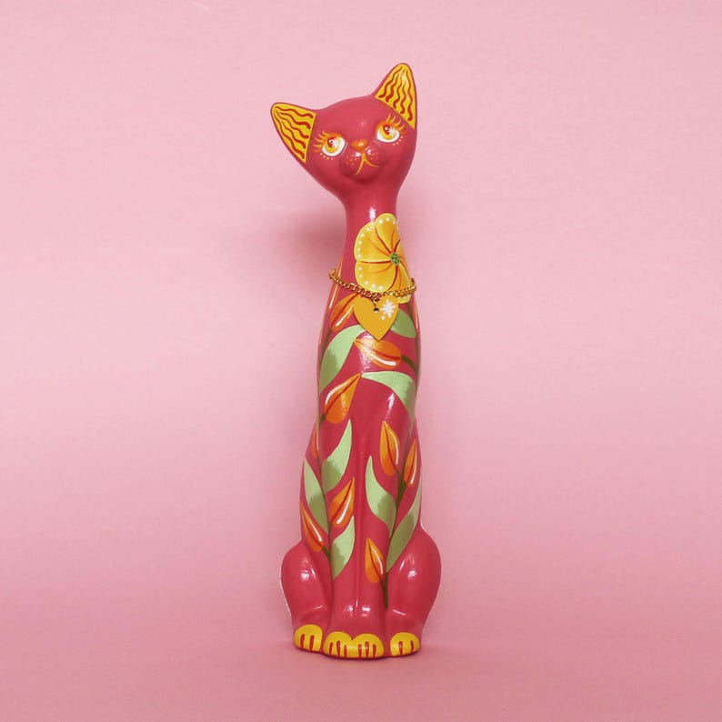 May include: A tall, pink cat figurine with yellow and green floral designs. The cat has yellow ears and a yellow flower necklace with a heart pendant. The cat's paws are yellow, and the background is pink.