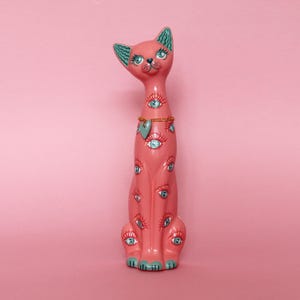 May include: A pink ceramic cat figurine with multiple blue-eyed designs. The cat has a gold chain necklace with a heart pendant. The ears and paws are teal. The cat is sitting on a pink background.