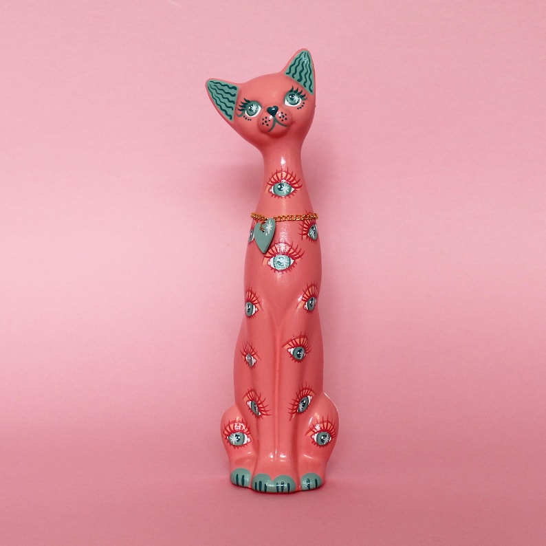 May include: A pink ceramic cat figurine with multiple blue-eyed designs. The cat has a gold chain necklace with a heart pendant. The ears and paws are teal. The cat is sitting on a pink background.