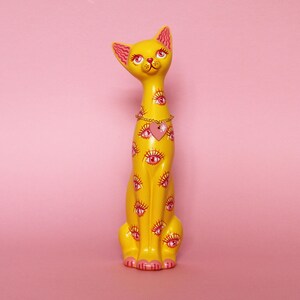 May include: A yellow cat figurine with pink accents, featuring multiple eye designs. The cat has a gold chain necklace with a pink heart pendant. The figurine is set against a pink background.