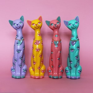 May include: Four colorful cat figurines in purple, yellow, pink, and teal. Each cat has a long neck, a heart pendant, and is decorated with multiple eye designs. The cats are posed sitting upright against a pink background.