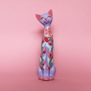 May include: A whimsical, lavender cat figurine with a long neck and upright posture. The cat is adorned with floral and heart motifs in red, green, and pink. It has a gold-colored necklace and is set against a pink background.