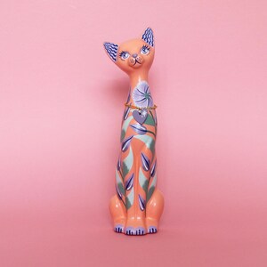 May include: A tall, decorative cat figurine in shades of peach, teal, and purple. The cat has blue eyes, patterned ears, and a floral design on its body. A gold-colored necklace with a heart pendant adorns its neck. The figurine is set against a pink background.