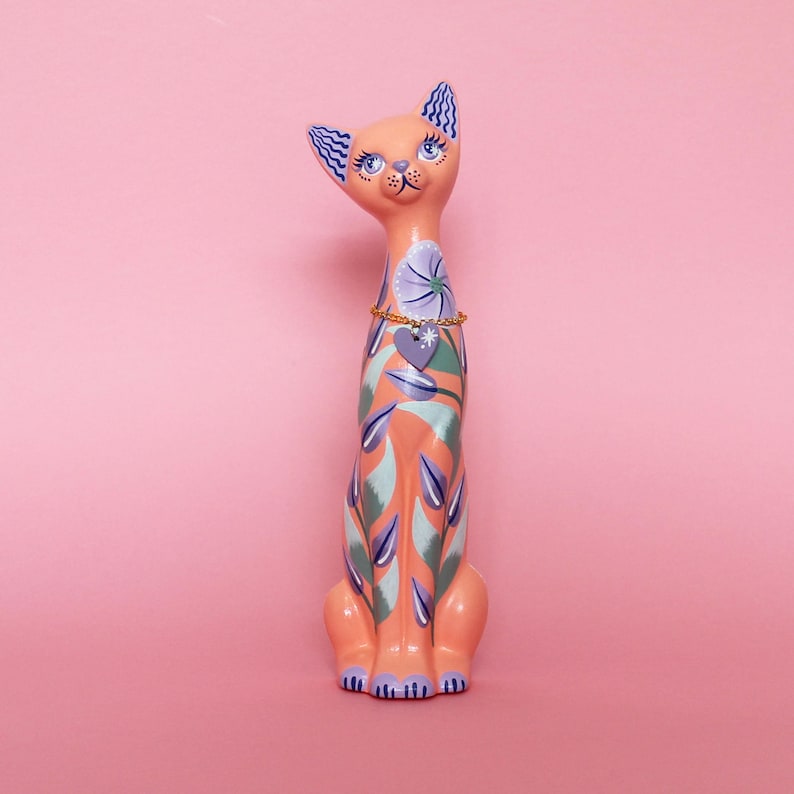 May include: A tall, decorative cat figurine in shades of peach, teal, and purple. The cat has blue eyes, patterned ears, and a floral design on its body. A gold-colored necklace with a heart pendant adorns its neck. The figurine is set against a pink background.