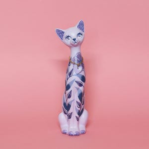 May include: A lavender ceramic cat figurine with blue and white floral and leaf patterns. The cat has blue eyes and a gold chain necklace with a heart pendant. The background is a solid pink.