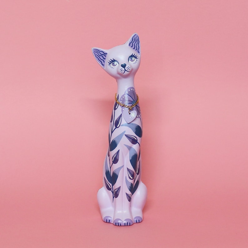 May include: A lavender ceramic cat figurine with blue and white floral and leaf patterns. The cat has blue eyes and a gold chain necklace with a heart pendant. The background is a solid pink.