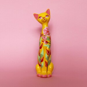 May include: A tall, yellow cat figurine with pink accents on the ears and paws. The cat is decorated with a floral design in red, green, and pink. A gold-colored chain necklace with a pink heart pendant adorns the cat's neck.