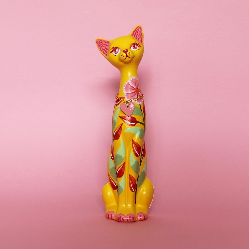 May include: A tall, yellow cat figurine with pink accents on the ears and paws. The cat is decorated with a floral design in red, green, and pink. A gold-colored chain necklace with a pink heart pendant adorns the cat's neck.