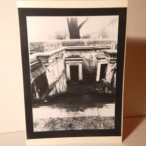 May include: Black and white photograph of a stone building with two doorways. The building has a weathered, aged appearance. The photograph is taken from a low angle, looking up at the building.