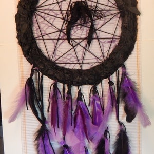Può includere: Un acchiappasogni fatto a mano con un anello nero strutturato e una ragnatela di filo nero. Presenta piume nere e viola appese dal basso, con nastri neri e dettagli in pizzo. Un pezzo decorativo per l'arredamento della casa.