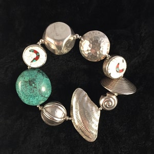 May include: A silver bracelet featuring a range of charms, including a turquoise stone, a white charm with red and blue accents, and a silver charm with a hammered finish.