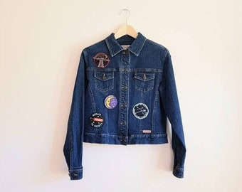 Peut inclure: Veste en jean bleu foncé ornée de plusieurs écussons brodés. Les écussons représentent une échelle, un croissant de lune et un motif céleste. La veste a une fermeture boutonnée, deux poches poitrine et des manches longues.