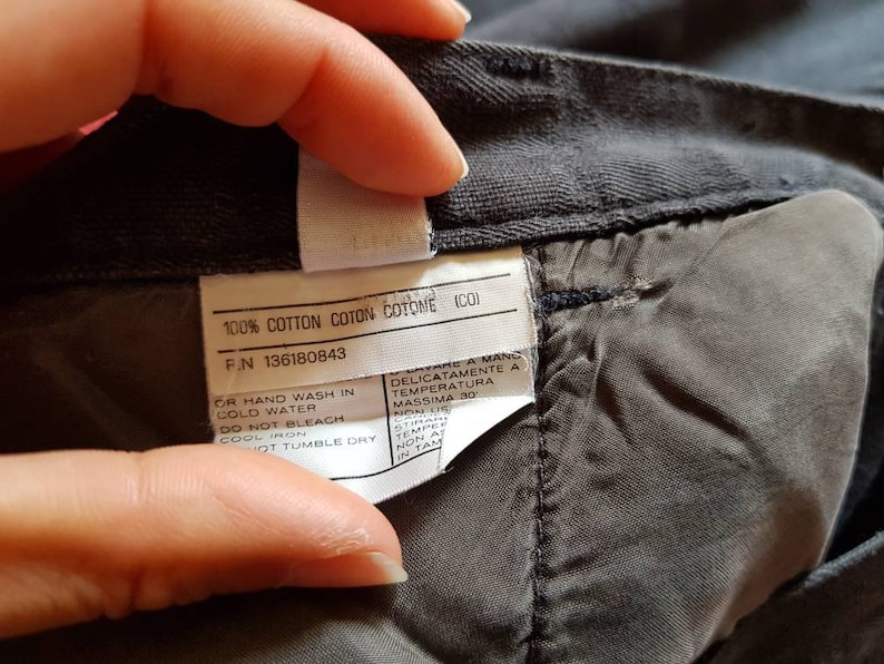 May include: A close-up of a grey cotton fabric label with care instructions. The label includes the text "100% Cotton Coton Cotome (CO)" and "F.N 136180843". The care instructions include "Or hand wash in cold water", "Do not bleach", "Cool iron", and "Hot tumble dry".