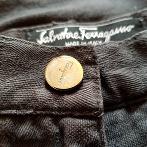 May include: Close-up of a dark grey pair of trousers with a gold button and a label that reads "Salvatore Ferragamo Made in Italy".