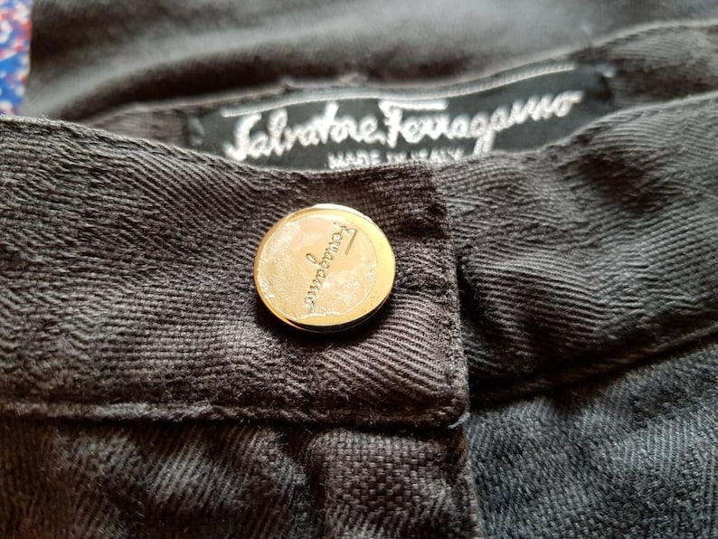 May include: Close-up of a dark grey pair of trousers with a gold button and a label that reads "Salvatore Ferragamo Made in Italy".