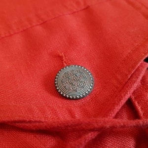 May include: A close-up of a red fabric jacket with a silver button featuring a decorative design. The jacket has a drawstring closure.