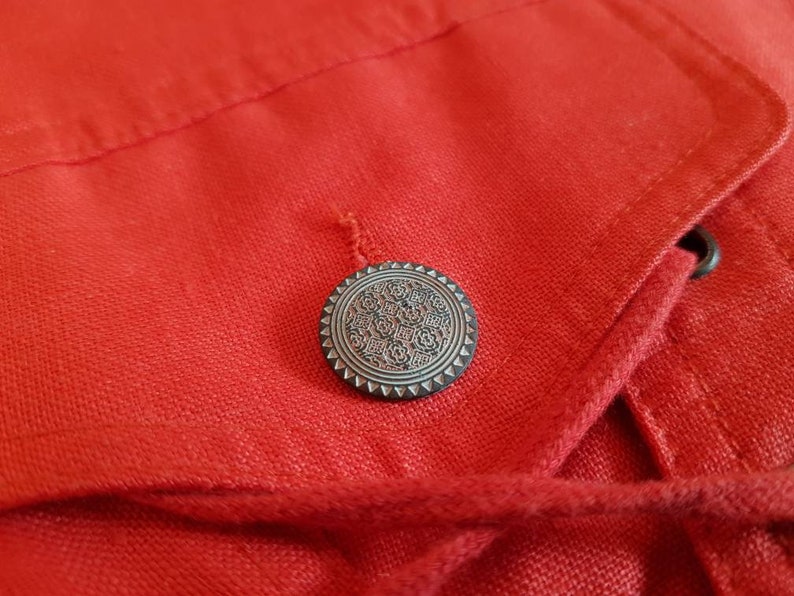 May include: A close-up of a red fabric jacket with a silver button featuring a decorative design. The jacket has a drawstring closure.