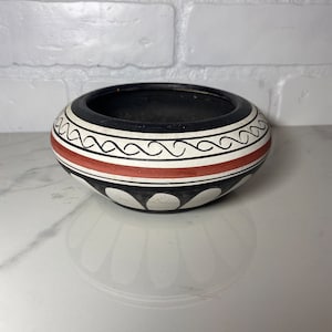 May include: A black and white ceramic bowl with a red band and geometric designs. The bowl is round and has a slightly flared rim.