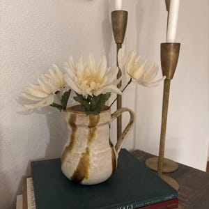 May include: A white ceramic pitcher with brown glaze sits on a stack of books. The pitcher is filled with white flowers and is next to a gold candle holder with three white candles.