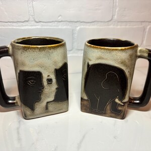 May include: Two ceramic mugs with brown handles. One mug features a black and white illustration of two faces. The other mug features a black and white illustration of an elephant.