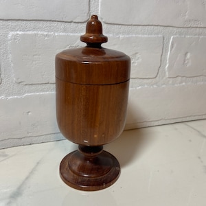 May include: A wooden box with a lid and a knob on top. The box is brown and has a pedestal base.