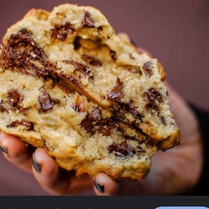 May include: A close-up of a chocolate chip cookie, broken in half. The cookie has a golden-brown exterior and is filled with large chunks of dark chocolate. The interior crumb is soft and chewy, with visible chocolate throughout.