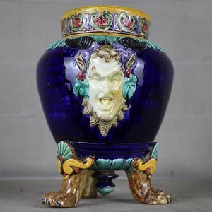 Puede incluir: Un jarrón de cerámica azul cobalto con un relieve de rostro blanco, sostenido por tres patas en forma de garra. El jarrón tiene un borde amarillo y con motivos florales. La base está sostenida por tres patas en forma de garra.