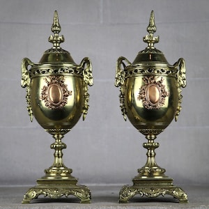 May include: A pair of ornate gold and copper urns with decorative details and a crest on the front. The urns are on a stone surface.