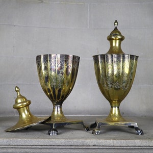 May include: Two antique silver and gold plated urns with ornate designs and a lid on a stone surface.