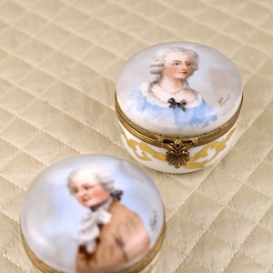 May include: Two small porcelain boxes featuring painted portraits of individuals in 18th-century attire. The boxes are round and have gold trim. Each box top is adorned with a floral design.