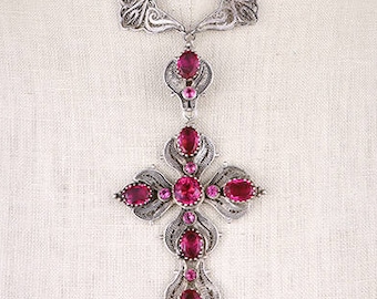 May include: A silver-toned pendant necklace featuring a cross design with pink gemstones. The necklace is displayed on a mannequin. The cross has an ornate filigree design and is suspended from a delicate chain.
