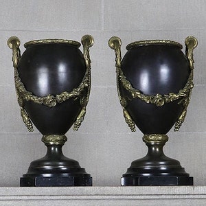 May include: A pair of black and gold decorative urns with ornate details. The urns are sitting on a stone mantelpiece.