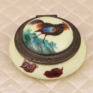 May include: A small, round, cream-colored porcelain trinket box with a hinged lid. The lid is decorated with a hand-painted image of a rooster in a landscape setting. The box is decorated with two small birds on the bottom.
