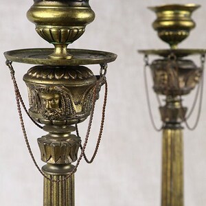 Puede incluir: Un par de candelabros de metal de color dorado, altos y con columnas estriadas, con detalles ornamentados. Cada candelabro tiene una parte superior decorativa con una cadena colgando.