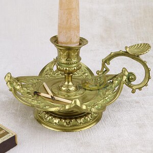 May include: A gold-toned brass candlestick with an ornate design. The candlestick has a single candle holder and a small tray for matches. The candle is a cream color and is partially burned.