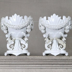 May include: A pair of white porcelain vases with intricate floral designs and a scalloped rim. The vases are sitting on a stone shelf.