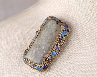 Pode incluir: Um broche retangular de jade com um design talhado, engastado em prata e decorado com esmalte azul e roxo. O broche é rodeado por um motivo floral.