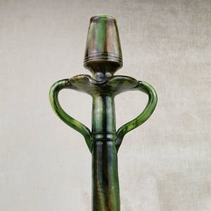 Pode incluir: Um castiçal de cerâmica verde e marrom com duas alças. O castiçal tem uma base larga e um caule alto e fino.