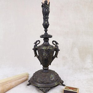 Puede incluir: Un candelero alto, ornamentado, de metal negro con una base decorativa y una cara esculpida en el centro. El candelero sostiene una vela larga y blanca.