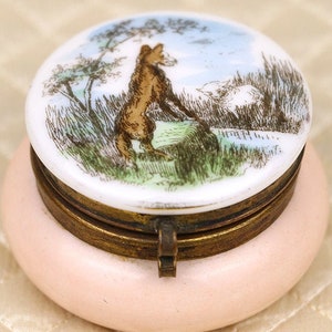 Puede incluir: Una pequeña caja de porcelana rosa redonda con tapa con bisagras. La tapa presenta una imagen pintada a mano de un zorro de pie en un campo de hierba con árboles en el fondo.