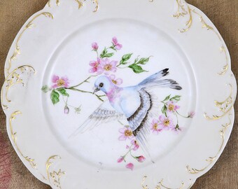 Pode incluir: Um prato de porcelana branca com uma borda dourada e um delicado desenho floral com um pássaro azul e rosa empoleirado em um galho com flores rosas.