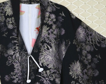 Könnte beinhalten: Ein schwarzer Seidenkimono mit silbernem Blumenmuster. Der Kimono hat eine weiße Schnur mit weißen Bommeln an den Enden. Der Kimono hängt an einem goldenen Haken.