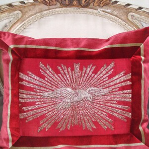 May include: A red velvet pillow with a gold embroidered design of a sunburst with a stylized bird in the center.