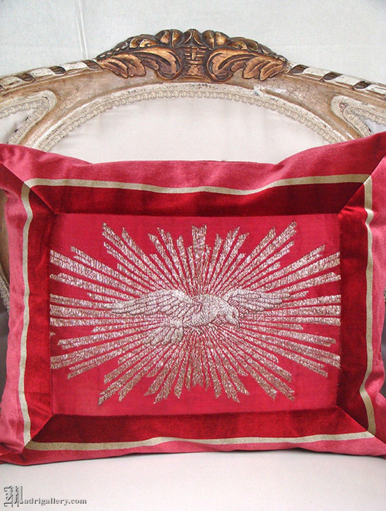May include: A red velvet pillow with a gold embroidered design of a sunburst with a stylized bird in the center.