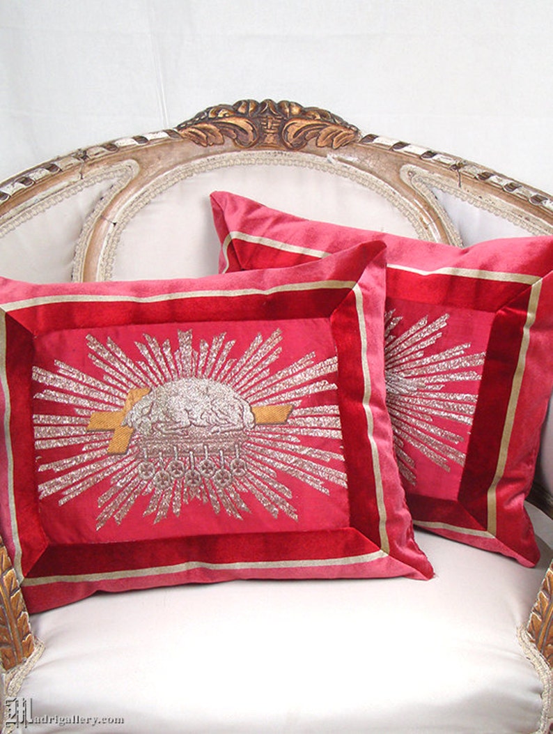 May include: Two red velvet throw pillows with gold and silver embroidery. The pillows feature a sunburst design with a central image of a crown.