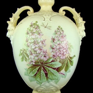 May include: A large, cream-coloured ceramic vase with two ornate handles in the shape of a dragon's head. The vase is decorated with a floral design of pink and white flowers and green leaves.