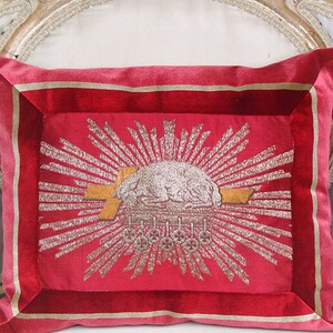 May include: A red velvet pillow with a gold and silver embroidered design of a sunburst with a cloud and a cross. The pillow has a gold and red border.