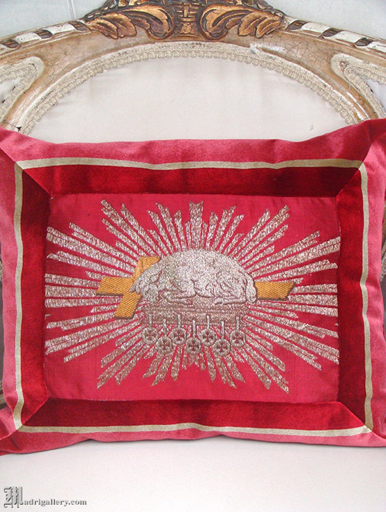 May include: A red velvet pillow with a gold and silver embroidered design of a sunburst with a cloud and a cross. The pillow has a gold and red border.