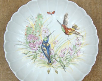 Może przedstawiać: Biały ceramiczny talerz z falistym brzegiem przedstawia ręcznie malowany wzór z dwoma kolibrami w locie, otoczonymi różowymi kwiatami i zielonymi liśćmi. Na talerzu przedstawiono również małego czerwonego motyla.