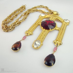 Puede incluir: Un collar de tono dorado con un gran colgante que presenta una piedra roja facetada rodeada de piedras más pequeñas transparentes. El colgante tiene forma de cruz con dos piedras colgantes en forma de lágrima.